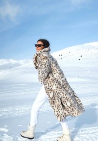 Leopárd mintás műszőrme kabát, hosszú és bolyhos, fehér nadrággal és bézs téli csizmával párosítva, havas táj előtt.