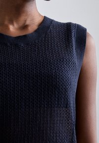 Gros plan d'une personne portant un haut sans manches en maille texturée bleu foncé avec un col rond, sur un fond uni clair.