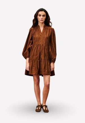 Femme debout portant une robe longue à manches longues en tiers marron et des chaussures Mary Jane marron sur un fond clair uni.