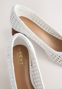 Witte gevlochten platte schoenen met een gesloten teen en een lage houten hak. Gestructureerd bovenwerk, glad interieur en een goudkleurig, geperst merklogo op de binnenzool.