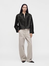 Woman with long straight hair wearing a black leather cropped jacket, beige wide-leg pants, and black pointed shoes standing against a plain background.
