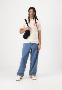 Beige grafisk t-skjorte med oransje trykk, blå vide bukser, og kremfargede sneakers med røde striper. Svart crossbody-veske med glidelåser.