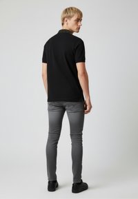 Černé polo tričko s kontrastním límečkem s potiskem, nošené s šedými úzkými džíny a černými botami. Model je zobrazen zezadu.