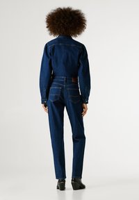 Džínová bunda a nohavice tmavomodrej farby. Bunda má golier a zapínanie na gombíky na manžetách. Nohavice majú zadné vrecká s koženým štítkom. Textúrovaná tkanina.