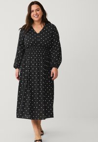 Schwarzes Kleid mit weißen Punktemuster, langen Ärmeln, V-Ausschnitt, geraffter Taille und fließender Silhouette, die bis zur Mitte der Wade reicht. Satinähnliche Textur.