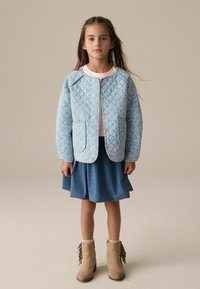 Giacca blu trapuntata con motivo floreale, due tasche e chiusura a bottoni a pressione, abbinata a una gonna di jeans e stivaletti alla caviglia beige con frange.