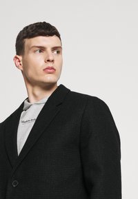Blazer negro a cuadros hecho de una tela texturizada con solapa de muesca y cierre de un botón, llevado sobre un sudadera gris.