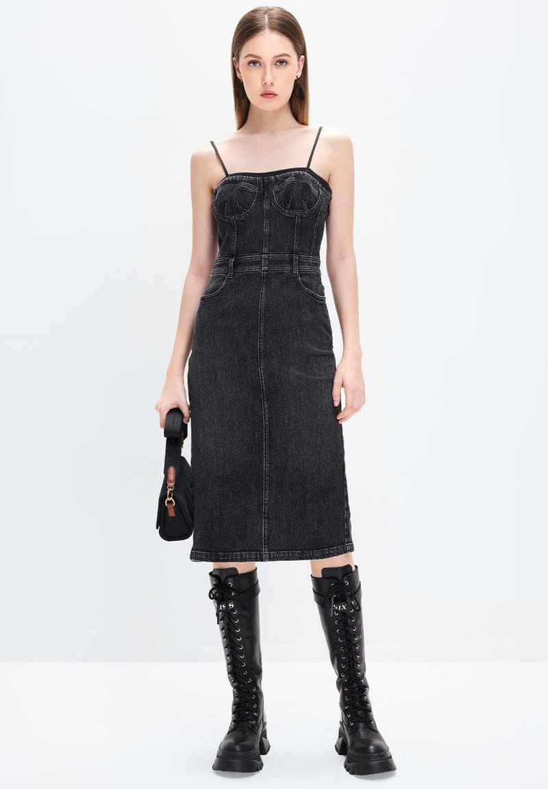 Vestido midi de denim negro con tirantes finos, corsé ajustado y bolsillos frontales. Combinado con botas altas negras de cordones. Diseño simple.