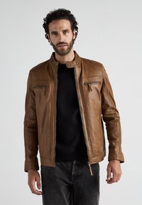 Mann mit lockigem Haar und Bart, der eine braune Lederjacke über einem schwarzen Shirt und dunklen Jeans trägt, steht vor einem schlichten Hintergrund.