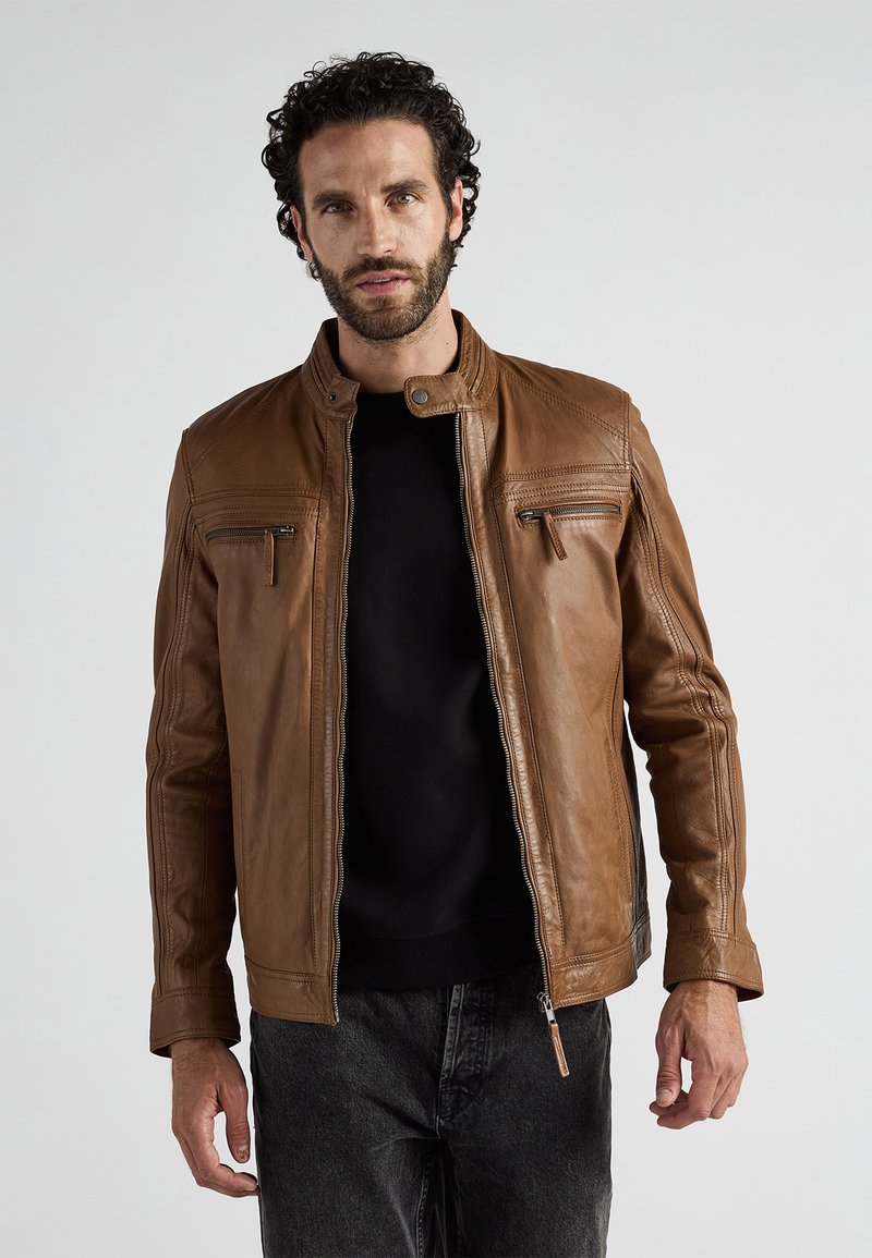 Mann mit lockigem Haar und Bart, der eine braune Lederjacke über einem schwarzen Shirt und dunklen Jeans trägt, steht vor einem schlichten Hintergrund.