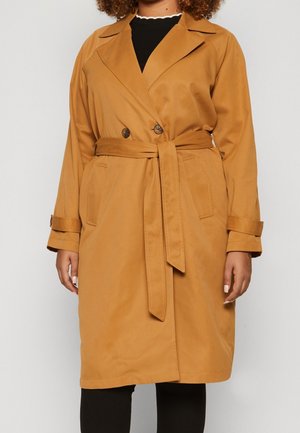 Kobieta w beżowym płaszczu trencz o długości do kolan, z paskiem w talii, dwurzędowymi guzikami i paskami na mankietach, nosząca czarne spodnie.