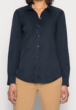 Femme portant une chemise bleu marine à manches longues boutonnée et un pantalon beige, debout avec les bras détendus le long du corps devant un fond uni.