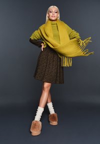 Maglione di lana verde oliva, indossato sopra una gonna marrone a fantasia. Sciarpa gialla con frange, calze color crema e pantofole foderate in pelliccia color cammello.