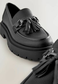 Mocasines negros hechos de cuero texturizado, con una suela de plataforma gruesa, borlas decorativas y acentos de metal.