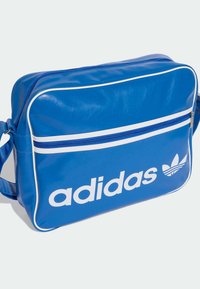 Geantă de messenger sintetică albastră cu accente albe, fermoar vizibil și un logo „adidas” alb proeminent, împreună cu trifoiul. Formă orizontală, textură netedă.