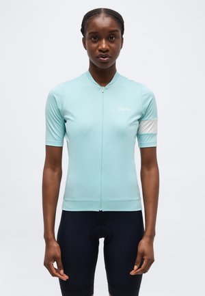 Femme portant un maillot de cyclisme à manches courtes bleu clair avec une bande blanche sur le bras et un short de cyclisme bleu marine, debout devant un fond uni.