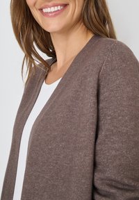 Cárdigan marrón de punto con diseño de escote en V, textura suave y abertura frontal. Llevado sobre una blusa blanca, con detalles de costura sutil.
