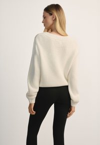 Kobieta o długich blond włosach ubrana w biały, gruby sweter z dzianiny i czarne dopasowane spodnie, stojąca tyłem.