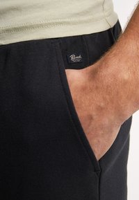 Pantalones de chándal negros con tejido texturizado, que presentan un bolsillo lateral y una pequeña etiqueta de logo cosida en la cintura. Una camiseta verde claro es parcialmente visible.