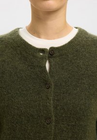 Grønn cardigan laget av mykt, lodden materiale, med rund halsutringning, knappestolpe foran med brune knapper, og en teksturert overflate.
