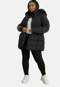 Svart pjäxjacka med huva av fuskpäls, kviltat mönster, två sidofickor, dragkedja framtill och elastiska ärmslut. Bärs över en vit topp och svarta leggings.