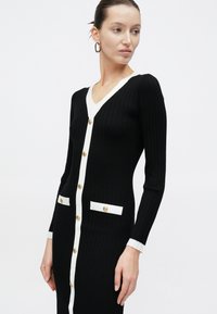 Γυναίκα που φοράει εφαρμοστό μαύρο ριμπ φόρεμα με λευκή επένδυση, χρυσά κουμπιά και δύο μπροστινές τσέπες, στέκεται μπροστά σε λευκό απλό φόντο.