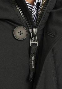 Chaqueta negra con cremallera, gran botón y tirador de marca. Presenta una textura suave y un forro a rayas en contraste.