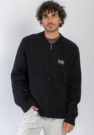 Hombre con el pelo rizado que lleva un jersey negro con cremallera con el texto "kaotiko society", de pie con una mano en el bolsillo de los pantalones claros.