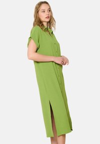 Abito camicia verde con bottoni, maniche corte, lunghezza al ginocchio, spacchi laterali, vestibilità rilassata, tessuto morbido e colletto.