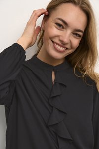 Χαμογελαστή γυναίκα με ανοιχτόχρωμο δέρμα και μπλε μάτια, που αγγίζει τα μαλλιά της, φορώντας μαύρη μπλούζα με ρυακιές μπροστά και μακριά μανίκια.