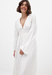 Hvid, langærmet kjole lavet af et struktureret stof. Har en dyb V-udskæring, snoreliv og en svævende silhuet.