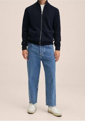 Maglione zip-up navy con polsini e orlo a costine, abbinato a jeans dritti azzurri e sneakers bianche con dettagli beige.