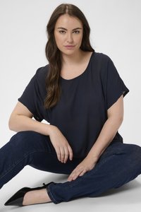 Námořnická modrá triko s krátkým rukávem a výstřihem do tvaru oválu, v kombinaci s tmavě modrými úzkými džíny a černými botami s špičatou špičkou, sedící pozice.