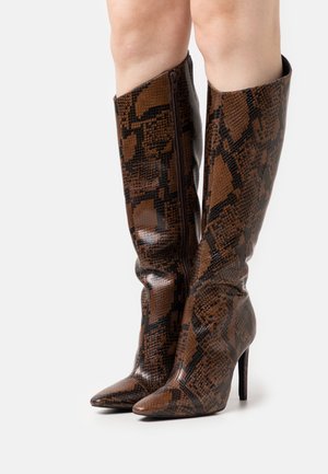 Femme portant des bottes à talons hauts montant jusqu'aux genoux, à bout pointu, à motif peau de serpent marron et noir, sur fond blanc.