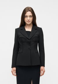 Γυναίκα με ίσια καστανά μαλλιά που φοράει ένα εφαρμοστό μαύρο σακάκι με δομημένες ραφές και δύο κουμπιά, στέκεται μπροστά σε απλό φόντο.