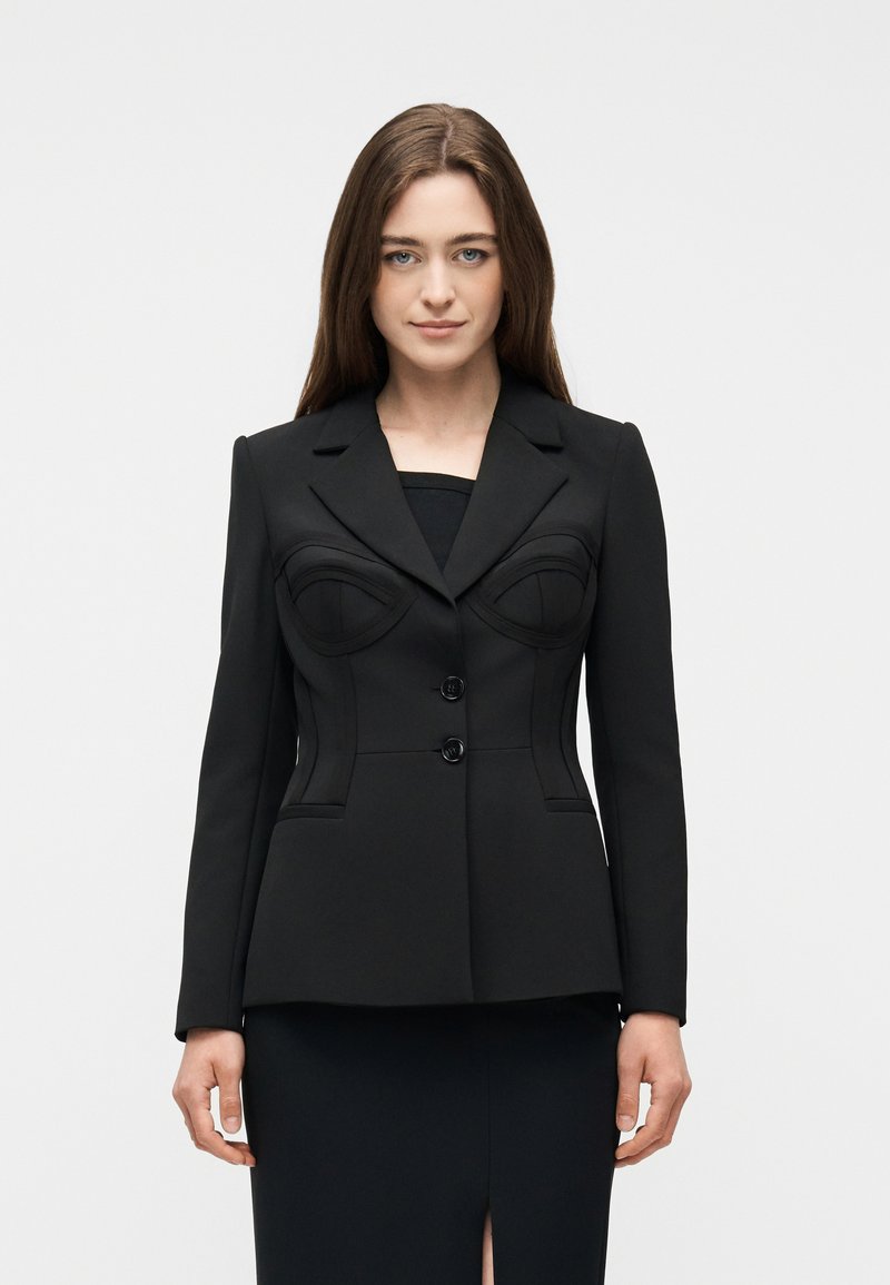 Γυναίκα με ίσια καστανά μαλλιά που φοράει ένα εφαρμοστό μαύρο σακάκι με δομημένες ραφές και δύο κουμπιά, στέκεται μπροστά σε απλό φόντο.