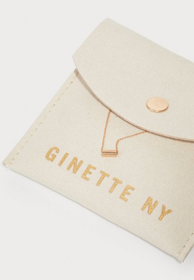 Vaaleanbeige huopa-pussi, jossa on läppäkiinnitys, kultaisesti painettu teksti "GINETTE NY" ja ruusukultainen ketjuyksityiskohta suorakulmaisen korosteen kanssa.