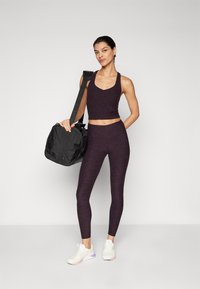 Mørk lilla treningscrop top og tights, med en svart duffelbag. Hvite sneakers med lilla og rosa detaljer. Glatt, tettsittende tekstur.