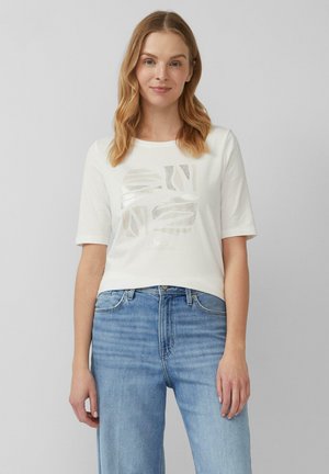T-shirt blanc à manches courtes avec un col rond et un design graphique métallique. Matière en coton léger ; coupe décontractée. Associé à un jean bleu.