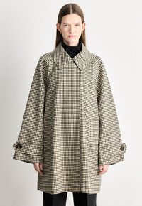 Manteau pied-de-poule marron et crème avec un grand col, des manches surdimensionnées et des accents à boutons. Deux poches avant visibles.