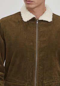 Ruda velveteen striukė su baltais dirbtiniais avikailio apykaklės kraštais ir priekine užtrauktuko uždarymo sistema. Pabrėžia vertikalią juostelę ir šonines kišenes.