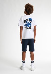 Witte t-shirt met kleurrijke grafische tekst "MIAMI FLORIDA" en blauw abstract ontwerp op de achterkant. Gecombineerd met marineblauwe shorts en witte sneakers.