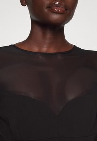 Robe noire avec un haut en maille transparente à encolure arrondie et un corsage ajusté. Texture lisse avec un léger éclat.