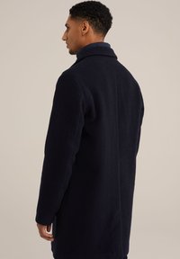 Marineblauwe wollen jas met een getailleerde snit, ingeknipte kraag en een gladde textuur. Zichtbare stiksels en lange mouwen met zijsplitten.