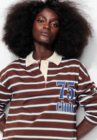 Polo rayé marron et blanc avec un col crème, présentant une broderie bleue "75 club" sur la poitrine gauche.