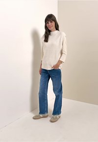 Jasno beżowy sweter z żebrowanym splotem i rękawami do łokcia, zestawiony z niebieskimi dżinsami i jasnobrązowymi sneakersami. Prosty design, gładka faktura.