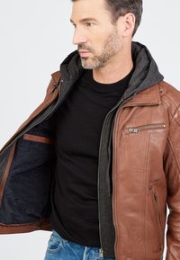 Ruskea nahkatakki harmaalla hupparin vuorella. Varustettu edessä vetoketjukiinnityksellä, sivutaskuilla ja sisätaskulla. Tasapintainen pinta, keskipitkä muotoilu.