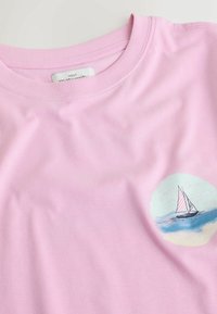 Gaiši rozā grūtnieču T-krekls ar ribotu apaļu apkakli un mazu burāšanas kuģa grafiku zilā, baltā un rozā krāsā uz krūtīm.