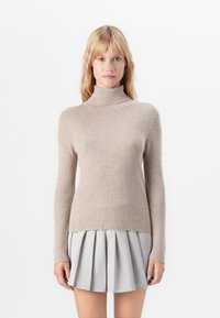 Kvinde iført en beige ribbestrikket turtleneck-sweater og en lysegrå plisseret mini-nederdel, stående foran en ensfarvet hvid baggrund.