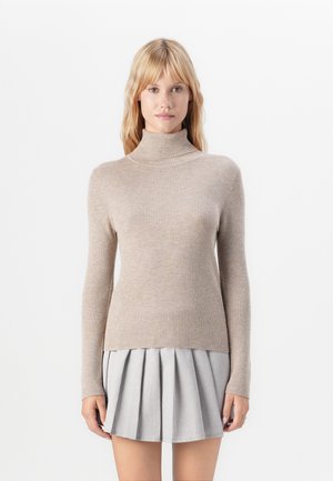 Kvinne iført beige ribbestrikket genser med høy hals og lysegrå, plisserte miniskjørt, stående mot en ensfarget hvit bakgrunn.
